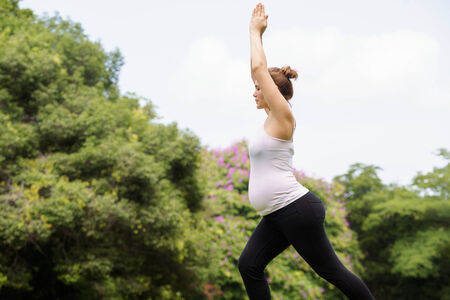 Portrait of white beautiful pregnant woman doing yoga exercise in park, with arms raised