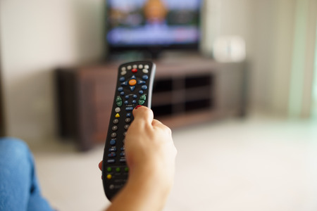 Over the shoulder view of girl sitting on sofa holding tv remote and surfing programs on television