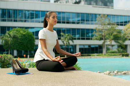 Young stressed hispanic business woman doing yoga outside office building, sitting in lotus position with hands on knees in the street. Concept of long working hours and need of stress free breakの写真素材