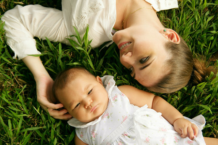 Young woman with her little daughter, relaxing on grass in park. The mom touches her toddler head...