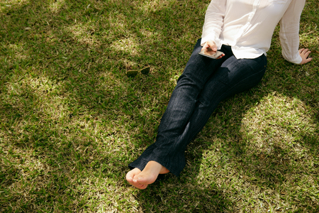 High angle view of young woman in jeans and white shirt resting in city park, sitting on grass and text messaging with mobile phone. Copy space, cropped viewの写真素材