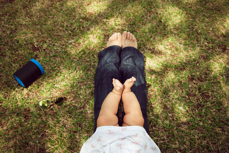 High angle view of little baby sitting on her mother legs on grass in city park. Concept of diversity, growth, contrast between small and big. Cropped view, copy spaceの写真素材