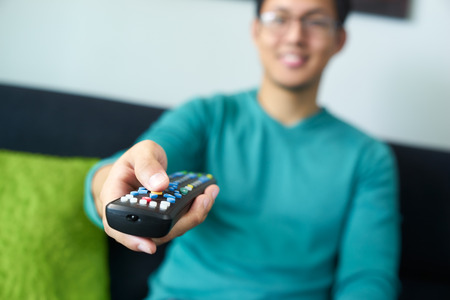 Asian young adult man watching TV and changing channel with remote control. Narrow focus on buttons and hand in foregroundの写真素材