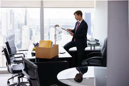 Businessman recently hired for corporate job moves into his new office. He takes out a folder from a box and reads documents near a big window, with a view of the city.の写真素材