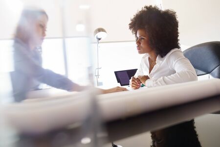 Team of two female architects, sitting at desk in office. The women talk reviewing a building plan. Medium shotの写真素材