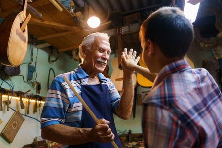 Small family business and traditions: old grandpa with grandson in lute maker shop. The senior artisan gives five to the boy, who helps him to make a handmade music instrumentの写真素材