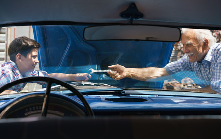 Family and Generation gap. Old grandpa spending time with his grandson. The senior man asks the preteen child to help him fixing the engine of a vintage car from the 60s. They smile happy. Viewed from the interior of the carの写真素材