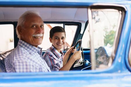 Family and Generation gap. Old grandpa spending time with his grandson. He teaches him to drive. The boy holds the volante of a vintage car from the 60s. They both smile happy looking at camera.の写真素材