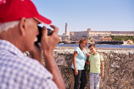 Happy tourists on holidays. Hispanic people traveling in Havana, Cuba. Grandfather, grandmother and grandchild during summer travel, with senior man taking photos with cameraの写真素材