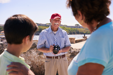 Happy tourists on holidays. Hispanic people traveling in Havana, Cuba. Grandfather, grandmother and grandchild during summer travel, with senior man taking photos with cameraの写真素材