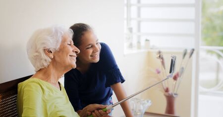 Family relationship between grandmother and granddaughter. Happy old woman and little girl at home taking self portrait picture with mobile phone and selfie stick.の写真素材