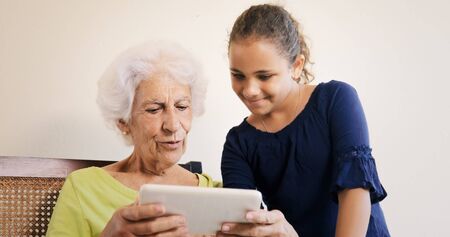 Family relationship between grandmother and granddaughter. Bond, affection and love, grandma and female grandchild surfing the Web with tablet at home.の写真素材
