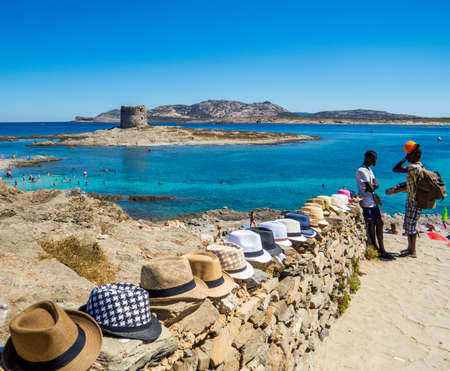 STINTINO, ITALY - JULY 20, 2016: African immigrants selling hats in front of the famous La Pelosa Beach with the landmark 16th century Watchtower (Italian: Torre della Pelosa) in the background.のeditorial素材