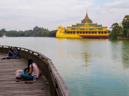 YANGON, MYANMAR - JANUARY 4, 2018: Local Burmese people on the wooden bridge over Kandawgyi Lake with the Karaweik Palace in the background.のeditorial素材