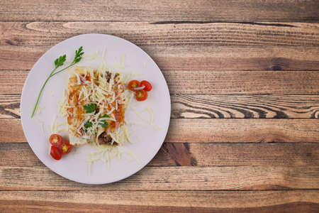 Pancakes with meat, in tomato sauce and cheese, with cilantro and grated cheese on top, and some cherry tomatoes on the plate. Top view on wooden background.の写真素材