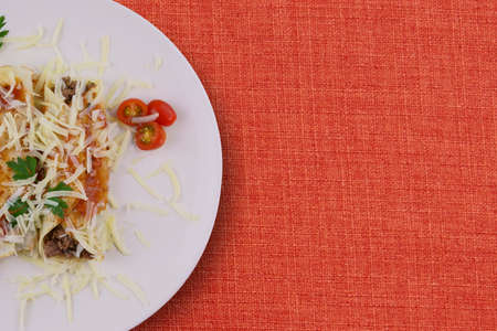 Pancakes with meat, in tomato sauce and cheese, with cilantro and grated cheese on top, and some cherry tomatoes on the plate. Top view on red background.の写真素材