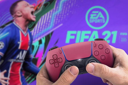Male hand holding a Playstation 5 Dual Sense Cosmic Red Controller with FIFA 21 game blurred in the...