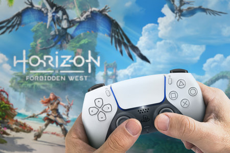 Male hand holding a Playstation 5 Dual Sense Controller with Horizon Forbidden West game blurred in...