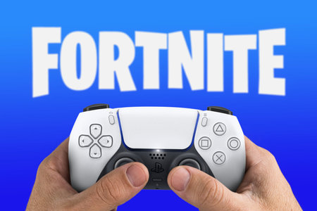 Male hand holding a Playstation 5 Dual Sense Controller with Fortnite game blurred in the...