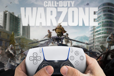 Male hand holding a Playstation 5 Dual Sense controller with Call of Duty Warzone game blurred in...