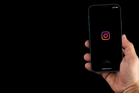 Male hand holding a smartphone with Instagram App on the screen. Social media. Black background. Rio...