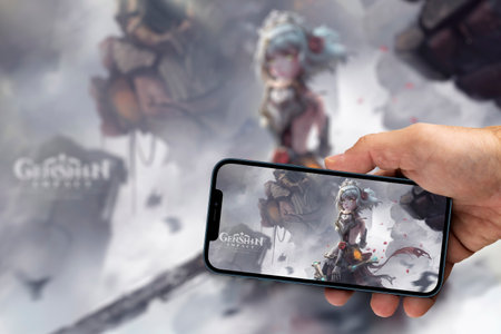 Male hand holding a smartphone with Genshin Impact game app on the smartphone screen with the game blurred in the background. Rio de Janeiro, RJ, Brazil. August 2021.のeditorial素材