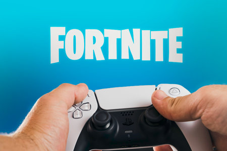 Male hand holding a Playstation 5 Dual Sense Controller with Fortnite game blurred in the...