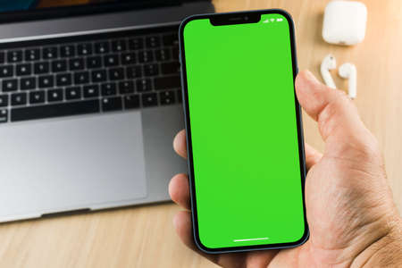 Male hand holding a smartphone with a green screen on wooden background with a computer. Top view. Chroma Key.の写真素材