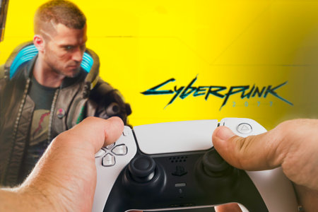 Male hand holding a Playstation 5 Dual Sense Controller with CyberPunk 2077 game blurred in the...