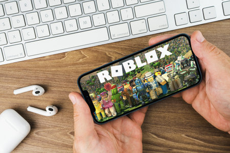 Male hand holding a smartphone with Roblox mobile game app on the screen with a computer keyboard beside it. Wooden Background. Rio de Janeiro, RJ, Brazil. October 2021.のeditorial素材