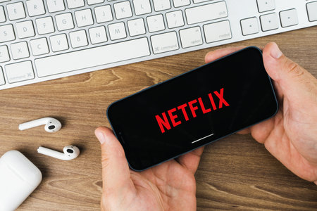 Male hand holding a smartphone with Netflix app on the screen with a computer keyboard. Wooden...