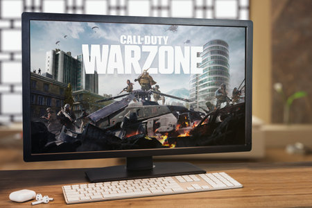 Call of Duty Warzone game on computer screen. Monitor, keyboard and airpods on wooden table....
