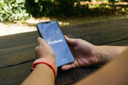 Girl in the park holding a smartphone with Facebook app on the screen. Rustic wooden table. Rio de Janeiro, RJ, Brazil. January 2022.のeditorial素材