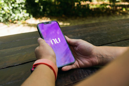 Girl in the park holding a smartphone with Nubank app on the screen. Rustic wooden table. Rio de Janeiro, RJ, Brazil. January 2022.のeditorial素材