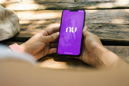 Girl in the park holding a smartphone with Nubank app on the screen. Rustic wooden table. Rio de Janeiro, RJ, Brazil. January 2022.のeditorial素材