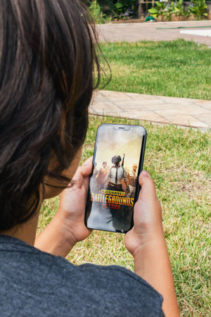 Child lying in hammock with Player's Unknown Battleground, PUBG mobile game app on smartphone screen. Lake in the background. Rio de Janeiro, RJ, Brazil. June 2022.のeditorial素材