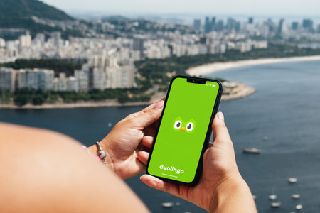Girl holding smartphone with Duolingo language-learning app on screen. City and bay with some boats in the background. Rio de Janeiro, RJ, Brazil. March 2022.のeditorial素材