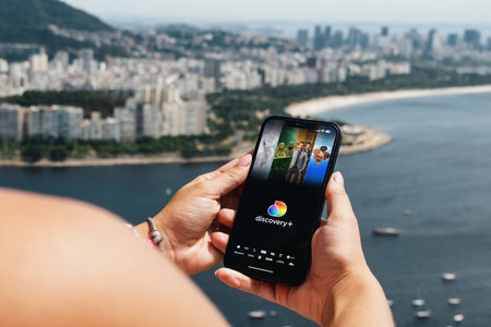 Girl holding smartphone with Discovery + (Plus) streaming app on screen. City and bay with some boats in the background. Rio de Janeiro, RJ, Brazil. July 2022.のeditorial素材