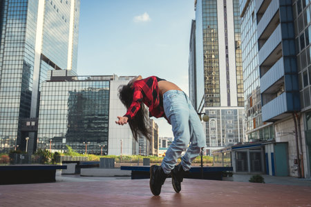 Photo shoot of stylish caucasian hip hop dancer posing on her toes with red shirt, jeans and black boots shoes and top with an urban town as background. City shooting of moving model bending the backの写真素材