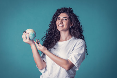 Studio shot of beautiful confident caucasian curly model woman wears blue jeans and casual basic solid white t-shirt, isolated over blue background. Pretty brunette holding small planet in her handsの写真素材