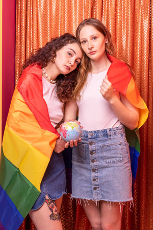 Attractive couple of casual caucasian girls posing standing with yellow - orange background hugging. Women with multicolor peace flag on shoulders holding the worldの写真素材