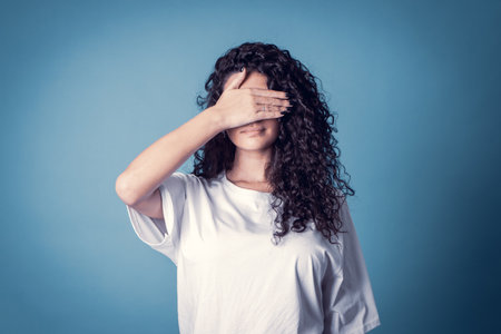 Shot of serious confident curly woman, wears casual basic solid white t-shirt, covering her face isolated over blue background. Expressionsの写真素材