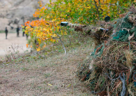 Camouflage Hunter or soldier aiming hiding in bushes in camouflage autumn  background. Sniper with rifle.の写真素材
