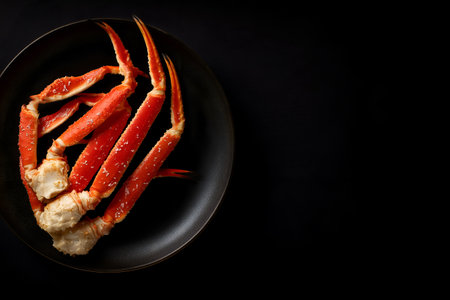 Black plate arranged with bright orange crab legs accented by white joints and scattered seasoning; dark background and directional lighting increase color contrast and texture, creating a dramatic, appetizing seafood presentation suitable for culinary, editorial, or menu photography.の素材