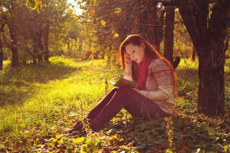 Young woman with long red hair reading under the tree in autumn forestの写真素材