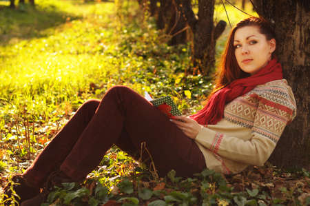Young woman with long red hair reading under the tree in autumn forestの写真素材