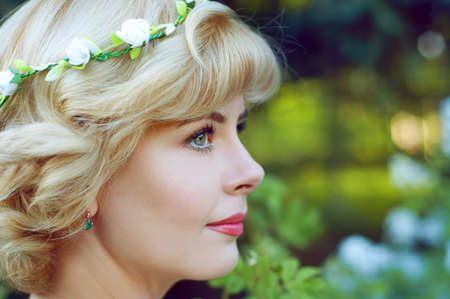Beautiful blonde woman in white sundress posing in garden wearing circlet of flowers