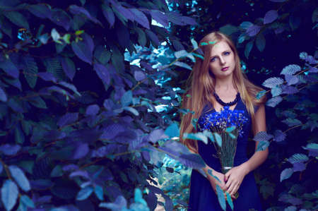 Blonde caucasian woman with long straight hair holding a cornflower bouquet posing in the labyrinth of treesの写真素材