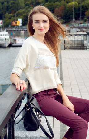 Tender young woman with long blond hair flowing on the wind sitting on the pier and smiling