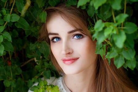 Happy smiling beautiful young woman surrounded by green leaves with blue eyes
